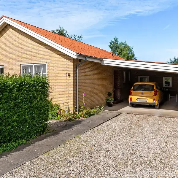 En gul murstenshus med rødt tag har en carport, hvor en gul bil er parkeret. Husnummeret "14" er synligt. Området er omgivet af buske og en grusindkørsel.
