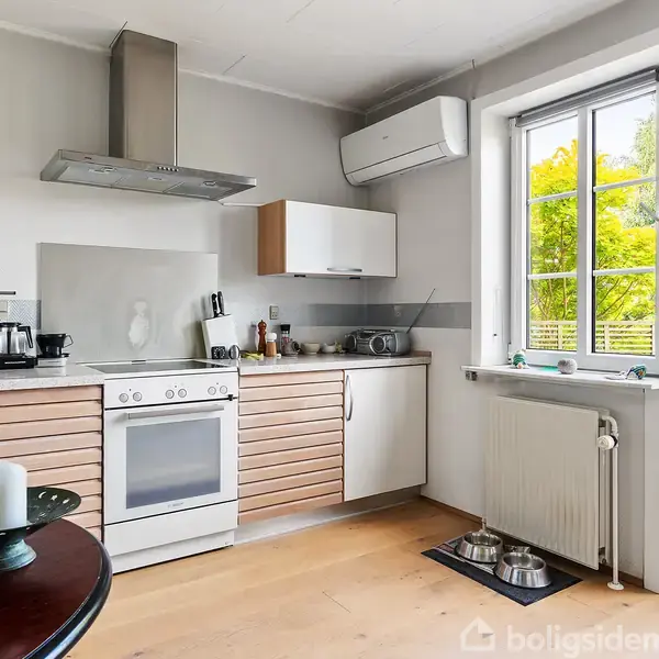 Et køkken indeholder et komfur med emhætte, køleskab og skab over en bordplade. Til højre er der vinduer og en dør med udsigt til en have. En radiator og hundeskåle er placeret ved døren. En rund spisebord med et lys i forgrunden.