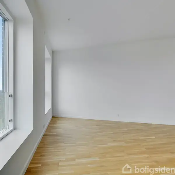 Et tomt rum med lyse trægulve har to store vinduer til venstre, der giver udsigt til byen. Væggene er hvide, og rummet er fyldt med naturligt lys.