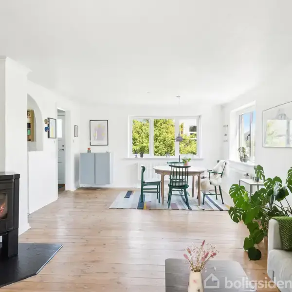 Pejs i stuen, brænder lyst, omringet af lyse trægulve. Ved siden står en sofa med grønne puder og en potteplante. Spisebord med stole ved vinduer.