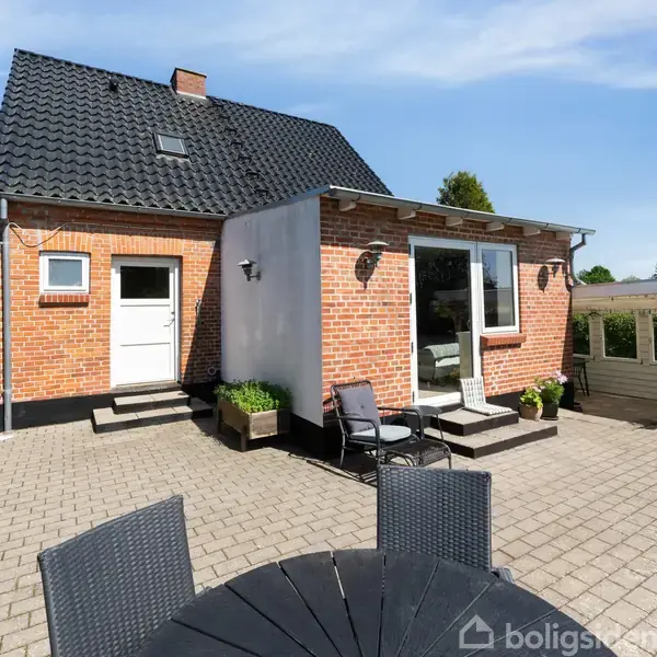 Et rødt murstenshus med sort tag og hvid dør står på en terrasse med fliser. Havemøbler og blomsterkrukker skaber en indbydende atmosfære under en klar blå himmel.