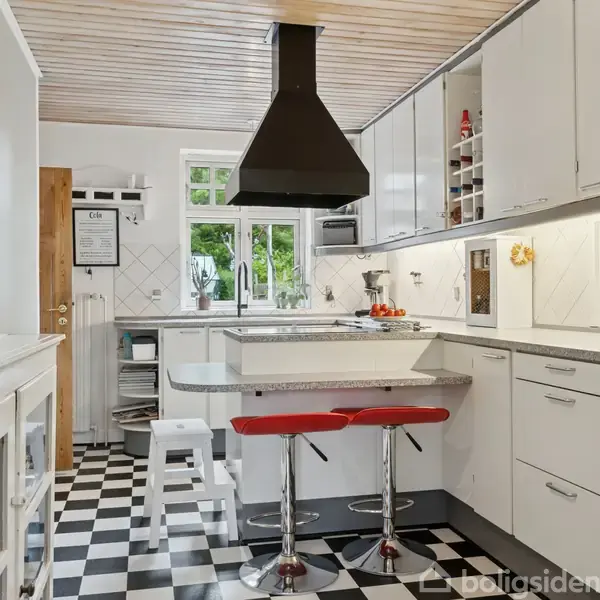 Køkken med hvide skabe og sort-hvidt skakternet gulv. To røde barstole står ved en central køkkenø under en stor sort emhætte. Vaser med blomster dekorerer området.