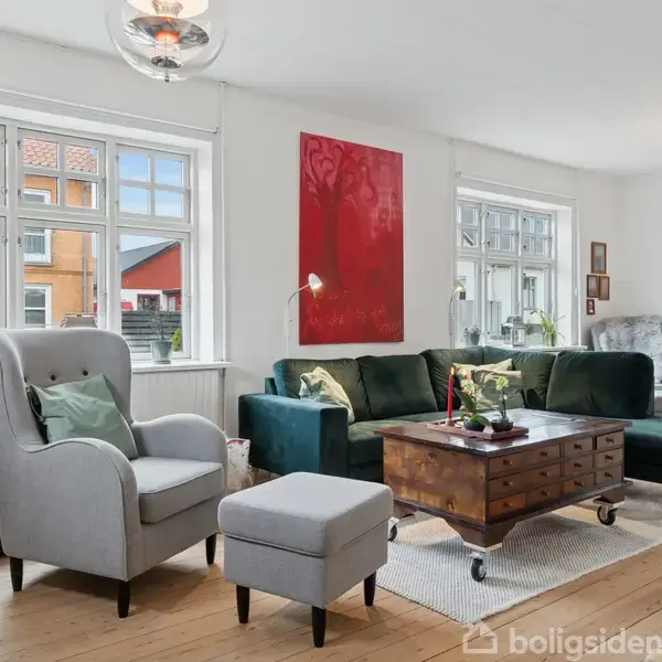 En lys stue med grøn sofa, lænestol og sofabord på hjul. Vægkunst i rød baggrund. To store vinduer bagved giver udsigt til nabobygning. Indbydende atmosfære med enkel indretning.