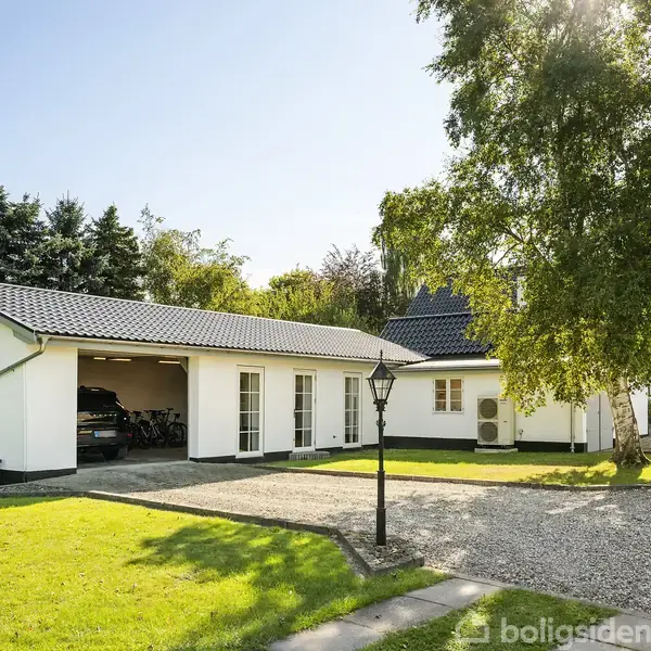 En hvid bygning med sort tag står ved siden af en grusvej. Garagen er åben med en bil indeni. Grøn græsplæne og træer omgiver huset.