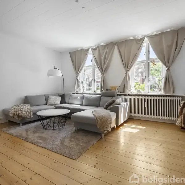 Grå sofa står i hjørnet af en lys stue med lyse trægulve. Ved siden af sofaen er et hvidt bord og en buet gulvlampe. Store vinduer med gardiner fylder væggen. En stol er dækket af tæpper ved siden af radiatoren.