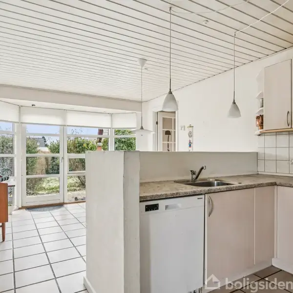 Køkken med hvide skabe og opvaskemaskine, oplyst af to pendellamper, åbner op mod stue med store vinduer og udsigt til have. Hvidt loft og flisegulv dominerer rummet.