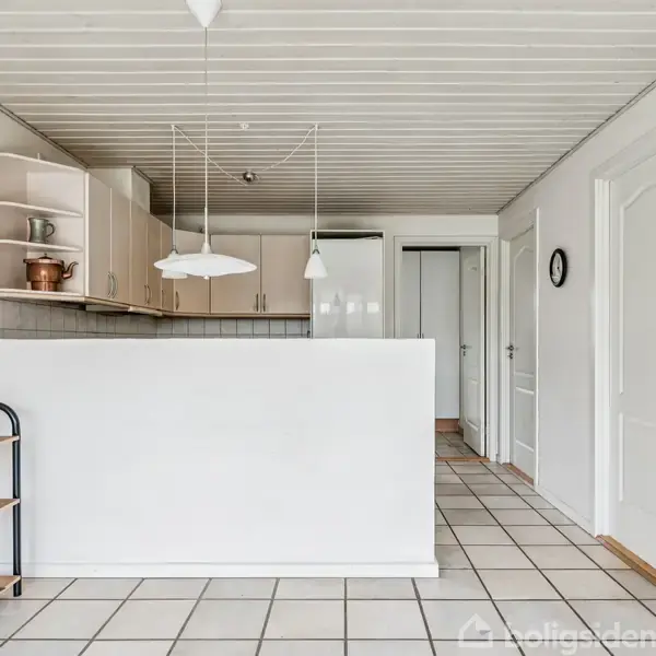 Et lyst køkken med hvide skabe og fliser, set fra stuen. Der hænger to lamper over en halv skillevæg. En åben reol står til venstre med dekorative genstande.