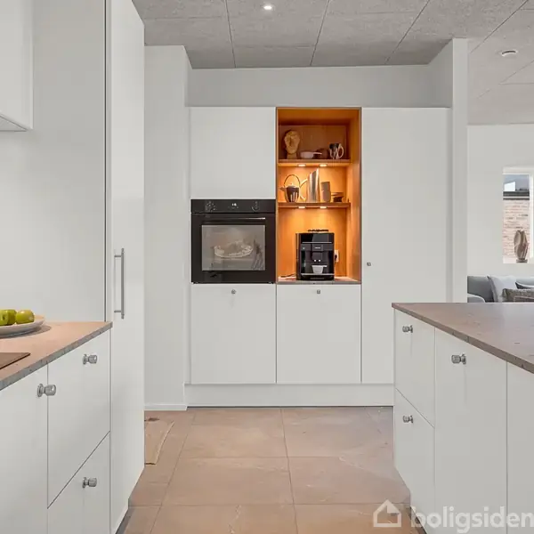 Et moderne køkken med hvide skabe indeholder en indbygget ovn og kaffemaskine. Skærebræt og krydderier står på bordet. Lys strømmer ind fra et vindue i baggrunden.
