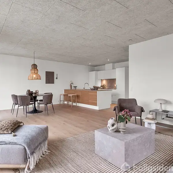 En moderne stue med minimalistik indretning har en grå sofa og et betonbord i forgrunden. I baggrunden ses et spisebord, en åben køkkenafdeling og flere grønne planter.