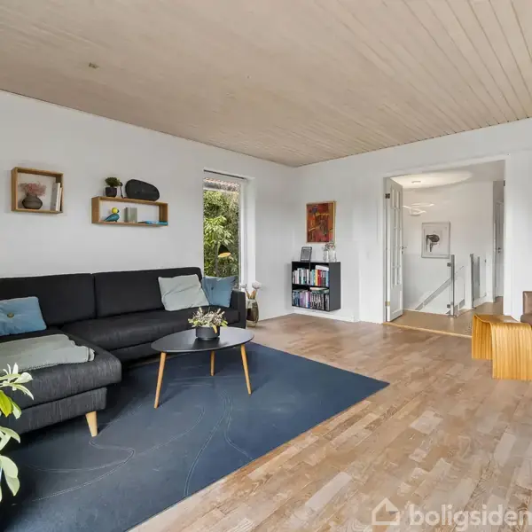 Sort sofa med puder står ved siden af et sofabord med planter i en stue med trægulv og hvide vægge. En døråbning fører til et andet rum.