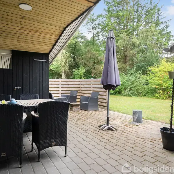 Terrasse med sorte kurvestole og bord. Parasol står foldet sammen. Omgivet af have med træer og grønne buske. Overdækket område til venstre, sort stakit i baggrunden.