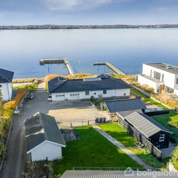 Hus ved vandområde står stille i kvarter med moderne bygninger og bådebroer, omgivet af græsplæner og parkeringsplads. Vandet strækker sig roligt ud under en grå himmel i baggrunden.