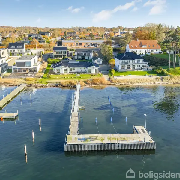 Mole med bådepladser strækker sig ud i vandet, omgivet af en rolig kystlinje og boligområde med huse og træer i efterårsfarver i baggrunden.