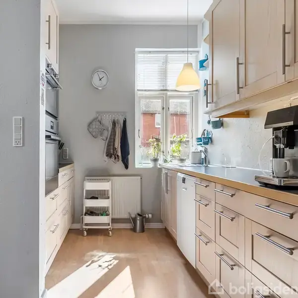 Et moderne køkken med lyse træskabe og en kaffemaskine på bordet. Et ur hænger over en lille reol med planter ved vinduet, gennem hvilket sollys strømmer ind.