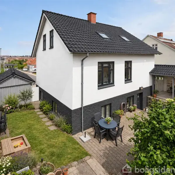 Et to-etagers hus med sort tag står midt i en velplejet have. En terrasse med rundt bord og stole foran huset. Nabobygninger ses i baggrunden. Tekst: "VISUALISERING".