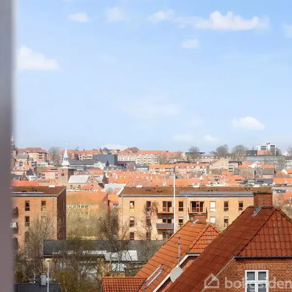 Udsigt over en by med røde tegltage og lejlighedsbygninger. I forgrunden ses et vindueshjørne, mens baggrunden viser flere boligblokke under en blå, skyfri himmel.