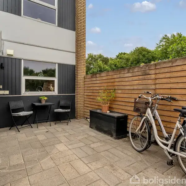 Cykel står parkeret på en flisebelagt terrasse ved siden af et hegn. To stole og et bord med en plante står ved husets facade.