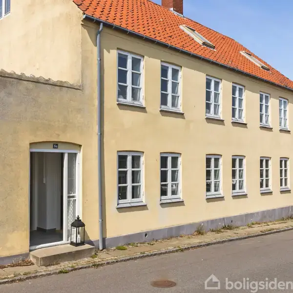 Et gult to-etagers hus med rødt tegltag står langs en vej. En lille lanterne er ved indgangsdøren. En rød bil er parkeret ved siden af.