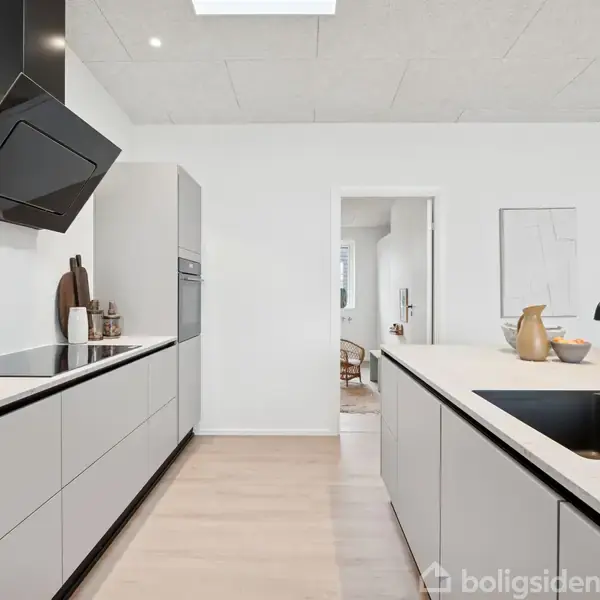 Moderne køkken med skabsflader og kogeplade, hvor emhætte hænger over. På den anden side er en vask indbygget i køkkenøen. Lyse vægge og trægulv skaber en minimalistisk atmosfære.