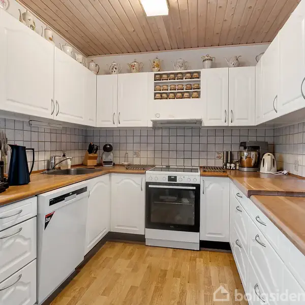 Et moderne køkken med hvide skabe og træbordplader. En ovn står centralt, flankeret af køkkenmaskiner. Rummet har trægulv og lyse lofter, med dekorative figurer oven på skabene.