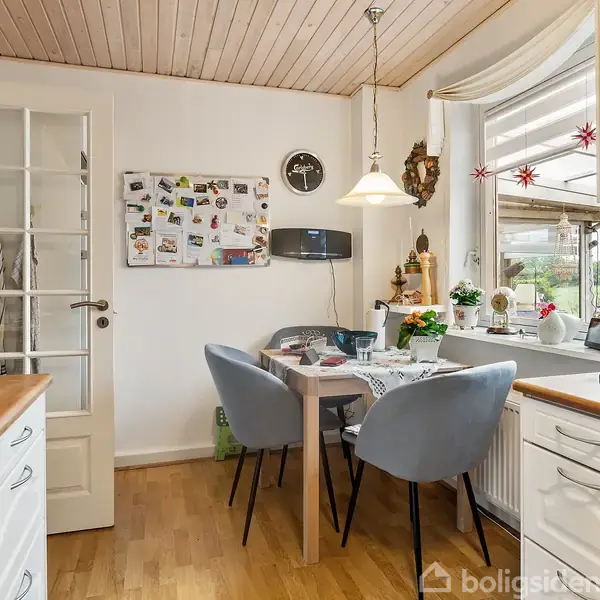 Et hyggeligt køkkenhjørne med et lille spisebord og fire grå stole. Bordet er dækket til måltid, omgivet af lyse vægge og et stort vindue med udsigt til haven.