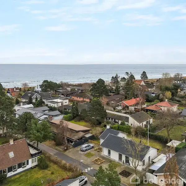 Huse med røde og sorte tage ligger tæt i et grønt kystsamfund. I baggrunden ses havet under en blå himmel med få skyer.