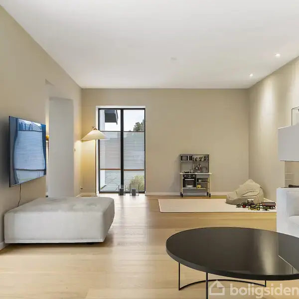 En stue med moderne indretning, stor fladskærms-TV på væggen, to sofaer, moderne sofabord, og gulvlampe bagest. Lyse trægulve og store vinduer danner rammen om rummet.