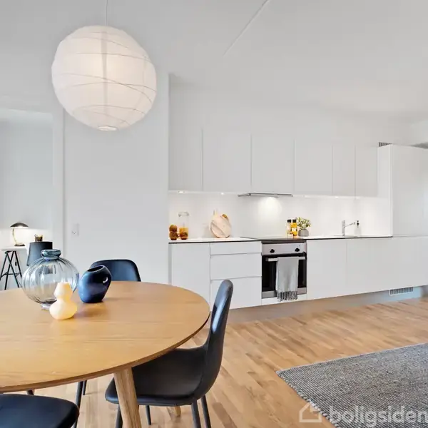Rundt spisebord med stole står i spisestue. I baggrunden ses et moderne køkken med hvide skabe og træparketgulv. En stor, rund loftlampe hænger over bordet.