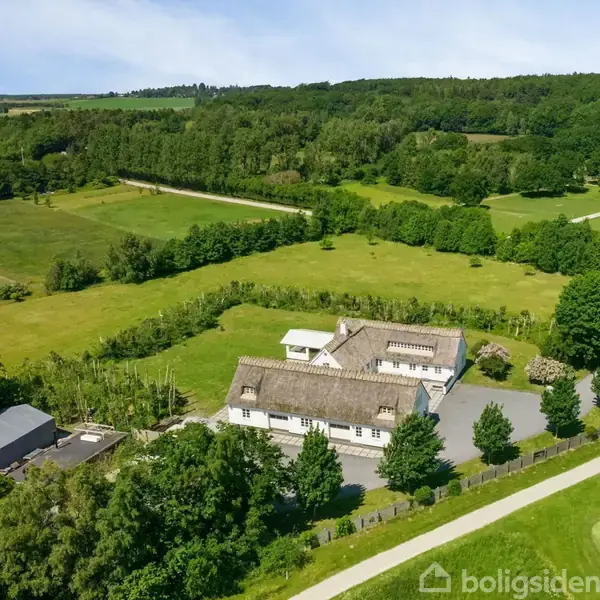 Hvide bygninger med gråt tag står stille i et grønt landskab med marker og træer omkring dem, under en klar blå himmel.