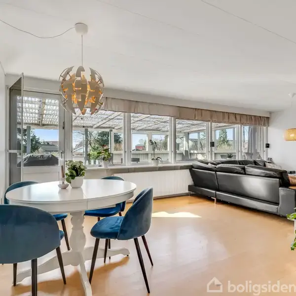 Rundt spisebord med blå stole står i et lyst rum med store vinduer. En sort hjørnesofa står ved væggen, og der er malerier og planter som dekoration.