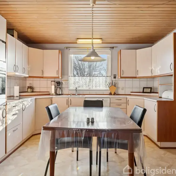 Et køkken med hvide skabe og trædetaljer. Et spisebord med fire stole står i centrum. En lampe hænger over bordet. Et stort vindue giver udsigt til haven.