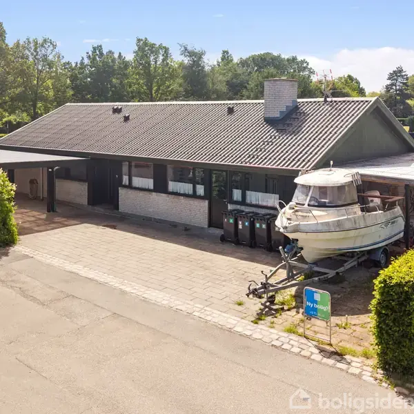 Et parcelhus med fladt tag har en båd på trailer foran. Husets indkørsel omgives af buske, og der er skov i baggrunden. Vejen ses i forgrunden.
