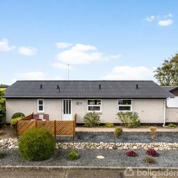 En hvid murstensbungalow med sorte tagsten står stille. En velplejet forhave med buske og sten omgiver huset. En delvis afskærmet terrasse er synlig ved indgangen.