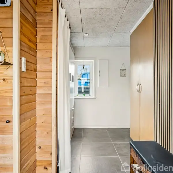 En smal gang med trægulv vægge, flisegulv, og en bænk til højre. Hylder med planter hænger ved en murstensvæg. Et vindue for enden giver udsigt til blå bygninger.