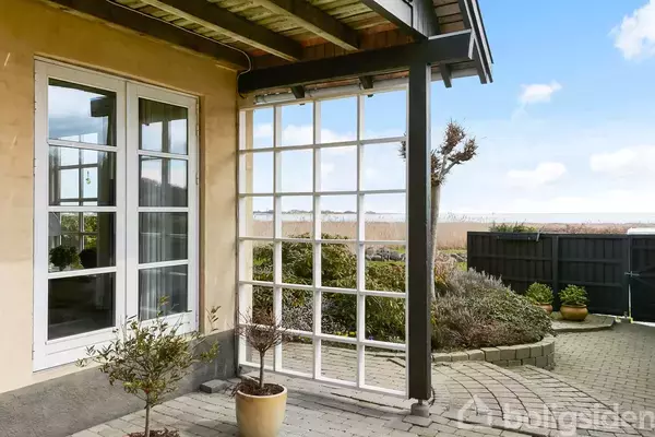 Et hus har store glasdøre, der åbner ud til en brolagt terrasse med blomsterpotter. I baggrunden ses en have og udsigt til havet under en blå himmel.