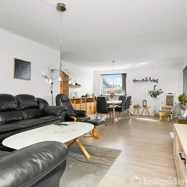 Sort lædersofa står ved et glasbord i stue med lyst trægulv. Spisebord med sorte stole ses i baggrunden. Rummet er lyst med store vinduer og grønne planter.