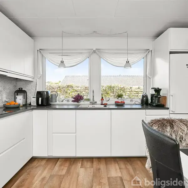 Hvidt køkken med moderne skabe og grå bordplade. Kaffeapparat og appelsiner står på bordet. Store vinduer med udsigt til have, pelsbetrukne stole forrest.