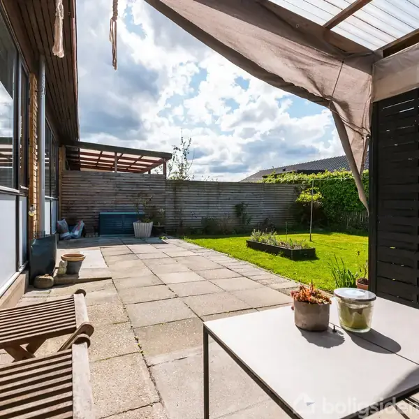 Træstole og et bord står på en flisebelagt terrasse under en halvtransparent overdækning. I baggrunden er der en grøn have med buske og en blå himmel med skyer.