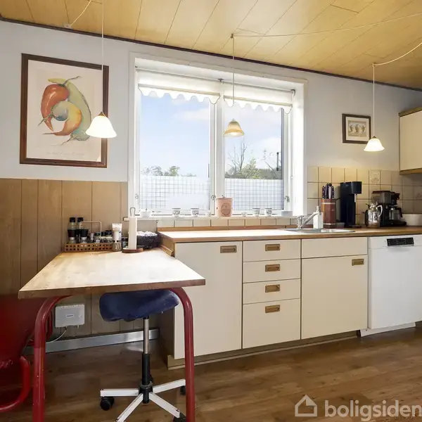 Et køkken med hvide skabe og brune håndtag omkranser et hjørnevindue. En bordplade støder op til en væg med rammet kunst. En rød bænk og stol står ved et spisebord.