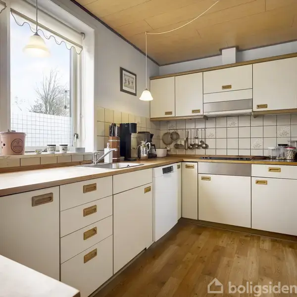 Et lyst køkken med hvide skabe og træbordplader. Der er en kogeplade og ovn integreret, køkkenredskaber hænger på væggen, og store vinduer slipper dagslys ind.