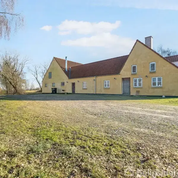 En gulmalet landejendom med rødt tegltag står under en blå himmel. Den er omgivet af åbne marker og træer, med en bred gårdsplads foran.