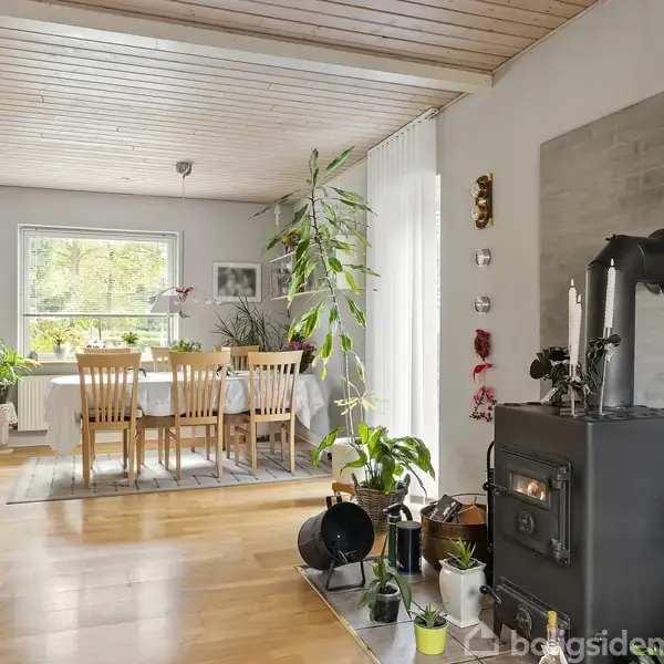 Brændeovn opvarmer en åben stue med trægulv og loft. Planteudsmykning omgiver rummet, hvor spisebord og stole er placeret ved et vindue, der giver naturligt lys.