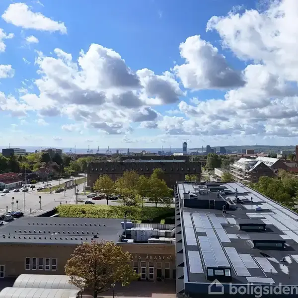 Bygninger med flade tage set fra en højde, omgivet af veje og træer under en blå himmel med spredte skyer. En fjern horisont med kraner og hav udsigt.