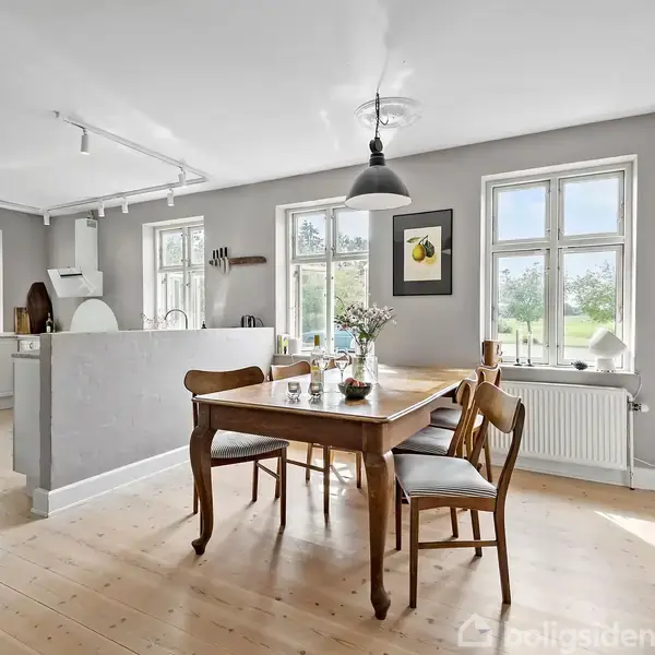 Et træspisebord står midt i et lyst køkken-alrum med hvidt loft og trægulv, omringet af stole og dekoreret med en blomstervase. Vinduet giver udsigt til grønne omgivelser.