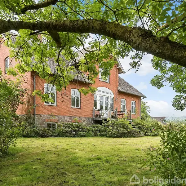 Et rødt murstenshus står stille i en frodig have under et stort træ. På facaden står der "DUGGEBAKKE." Græsset omkring huset er velplejet, og omgivelserne er grønne og rolige.