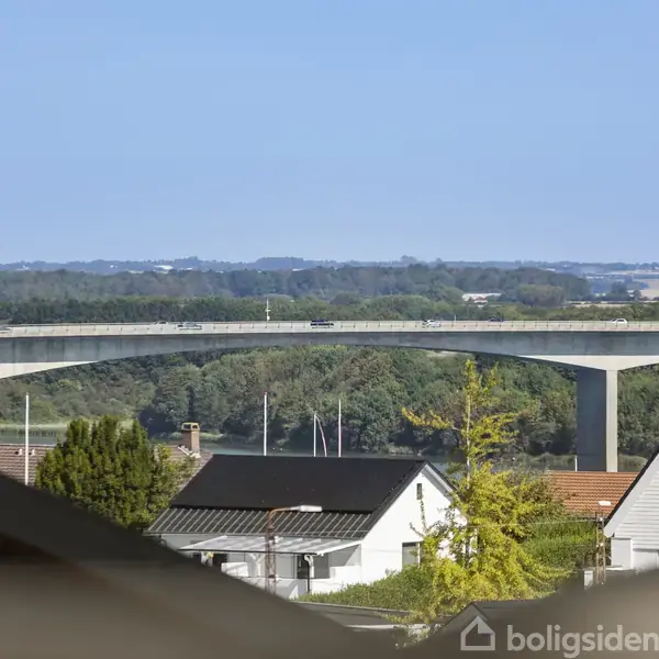En bro strækker sig horisontalt over en frodig dal. Foran ses hustage fra en forstad med træer og en klar blå himmel i baggrunden.