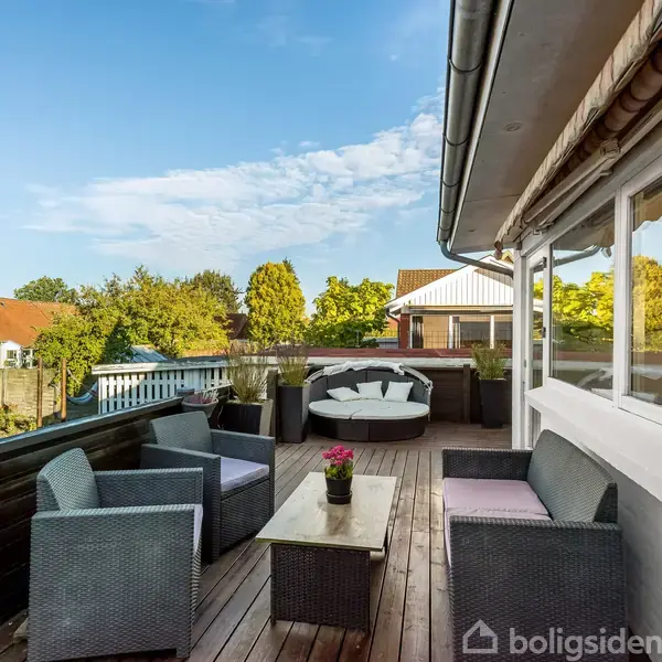 Terrassemøbler står på en træterrasse. Sofaer og et bord er arrangeret, mens himlen er klar. Omgivet af grønne træer og flere huse i baggrunden.