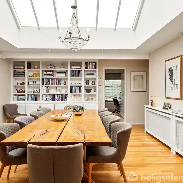 Træbord omgivet af polstrede stole står i spisestue med kunst på væggene. Bogreol fyldt med bøger i baggrunden, ovenlys tilfører rummet lys. Elegante detaljer skaber en moderne atmosfære.