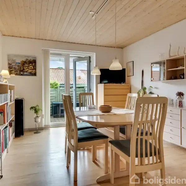 Et spisebord er omgivet af fire stole i et lyst rum. En dør med glasåbninger fører til en have. Hylder og en skænk fyldt med bøger og dekorationer står langs væggene.