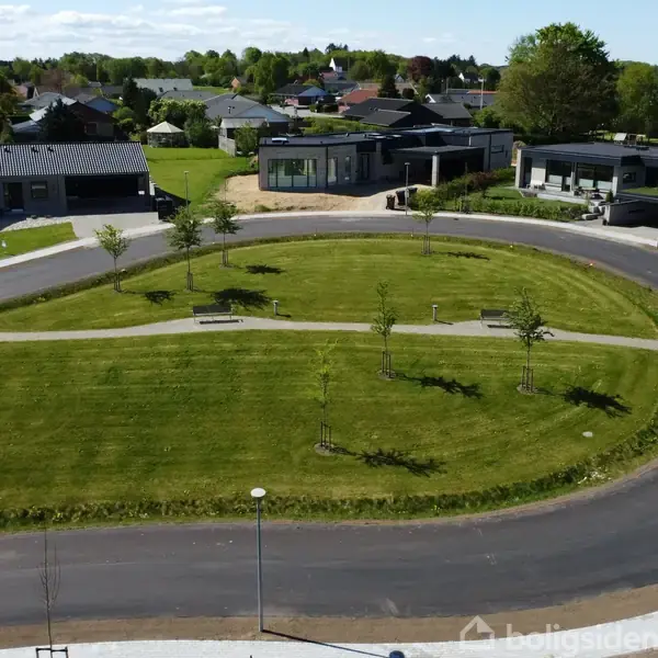 En velplejet græsplæne omkranset af en asfalteret vej, beliggende i et moderne boligområde med huse og træer i baggrunden. Der er bænke og unge træer på plænen.